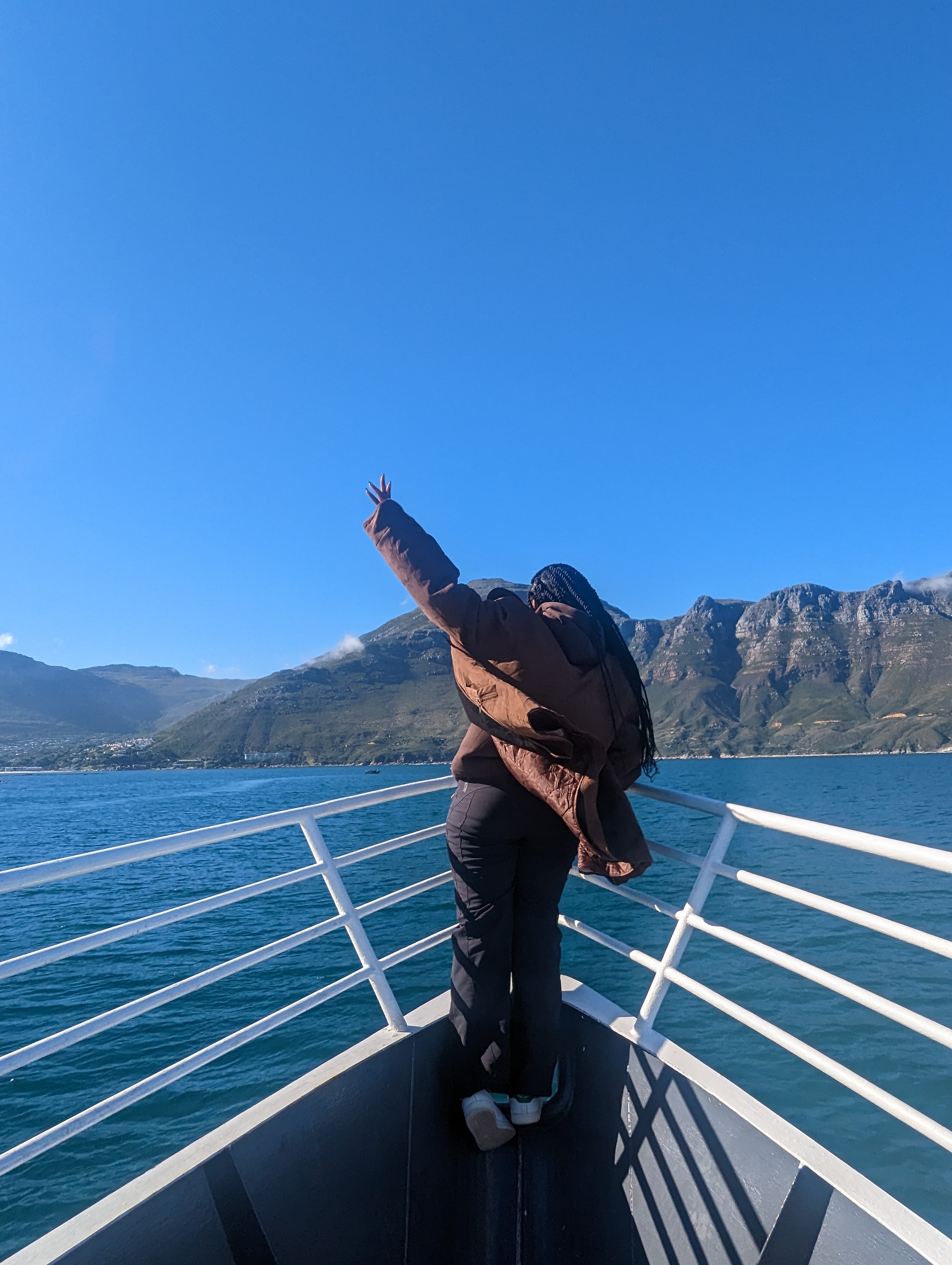Standing at the bow of a boat with one arm raised, mountains and open water ahead.