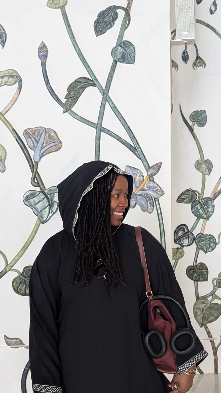 Liz in a hooded black abaya, looking off to the side and smiling, in front of a wall of painted leaves and flowers.