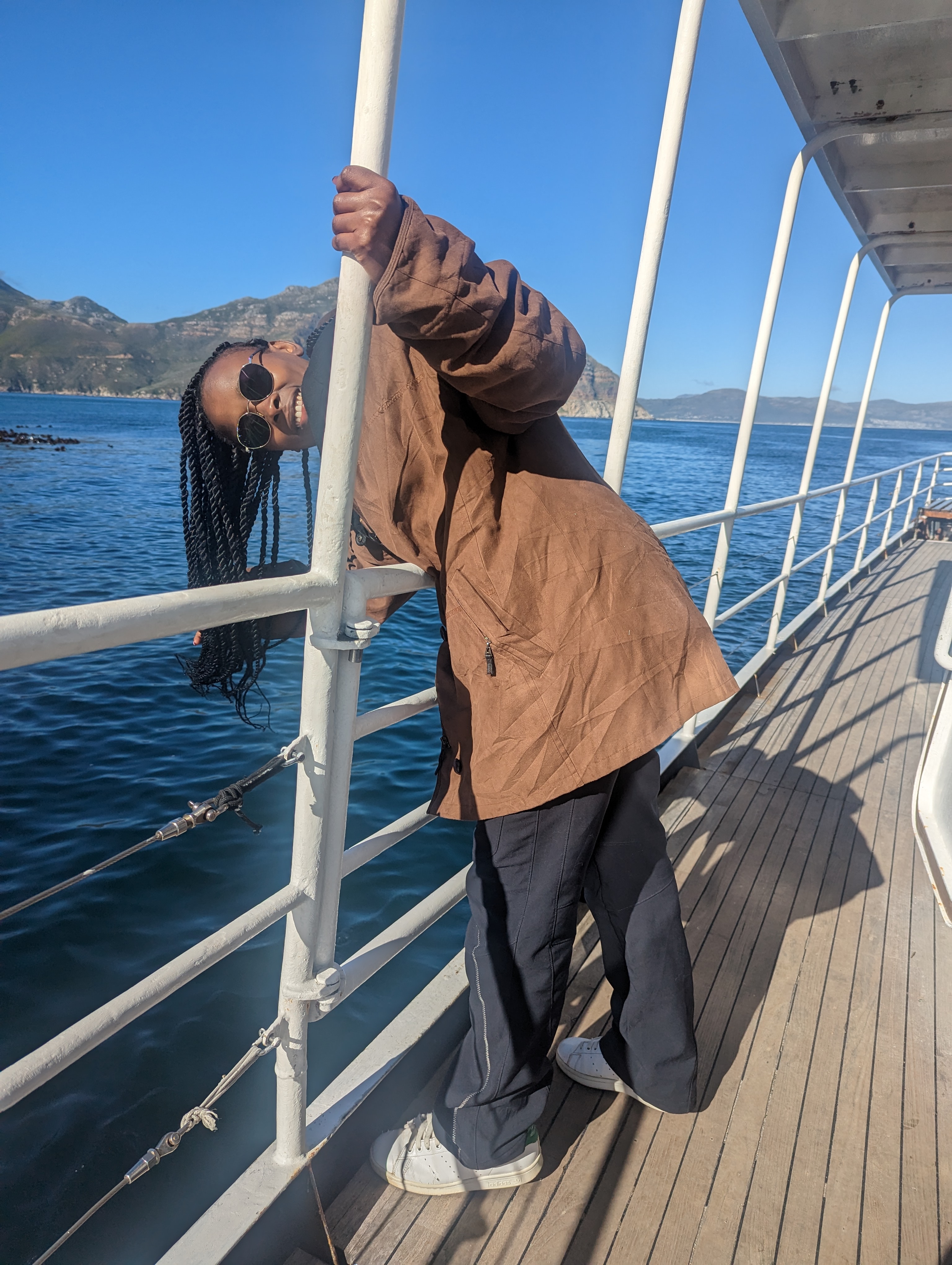 Leaning off the rail of a boat in a brown jacket and round sunglasses, laughing.